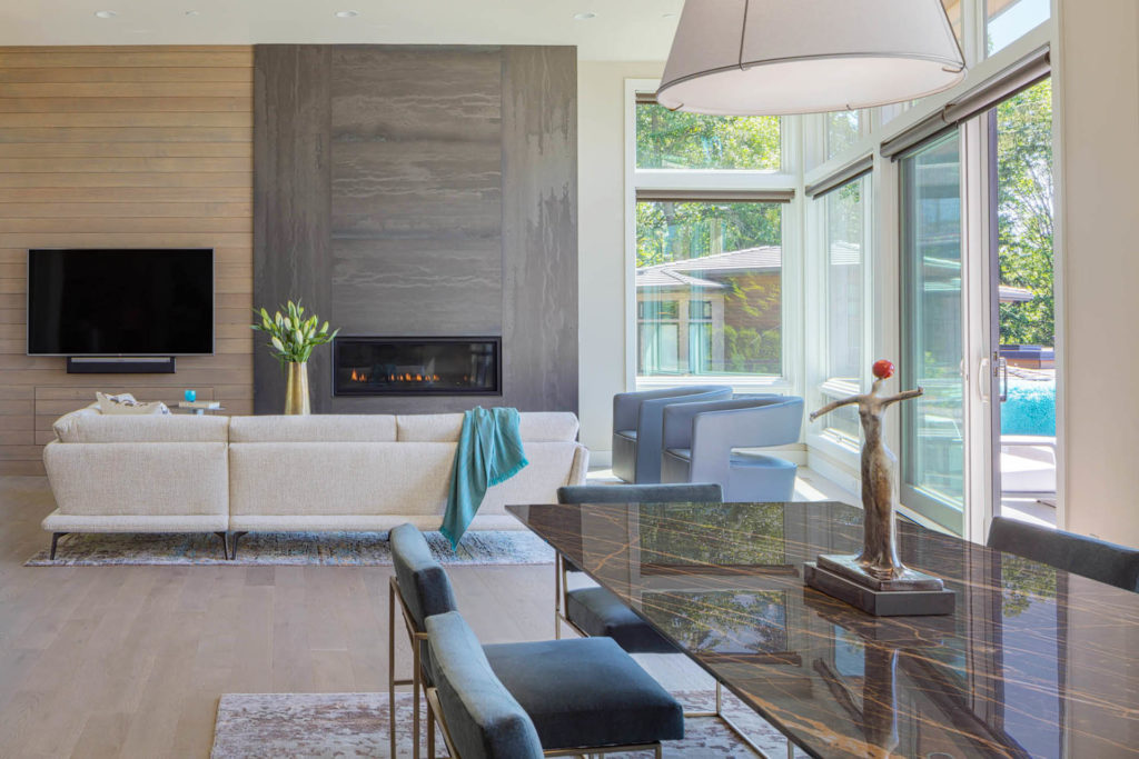 Modern open-concept living and dining area with large floor-to-ceiling windows letting in natural light. A beige sectional sofa faces a wall-mounted TV and a linear fireplace framed by a dramatic floor-to-ceiling hot-rolled steel surround, with a wooden accent wall on the left. A dark marble dining table with a bronze sculpture centerpiece and upholstered dining chairs sits in the foreground. The room features light wood floors and a neutral, contemporary palette.